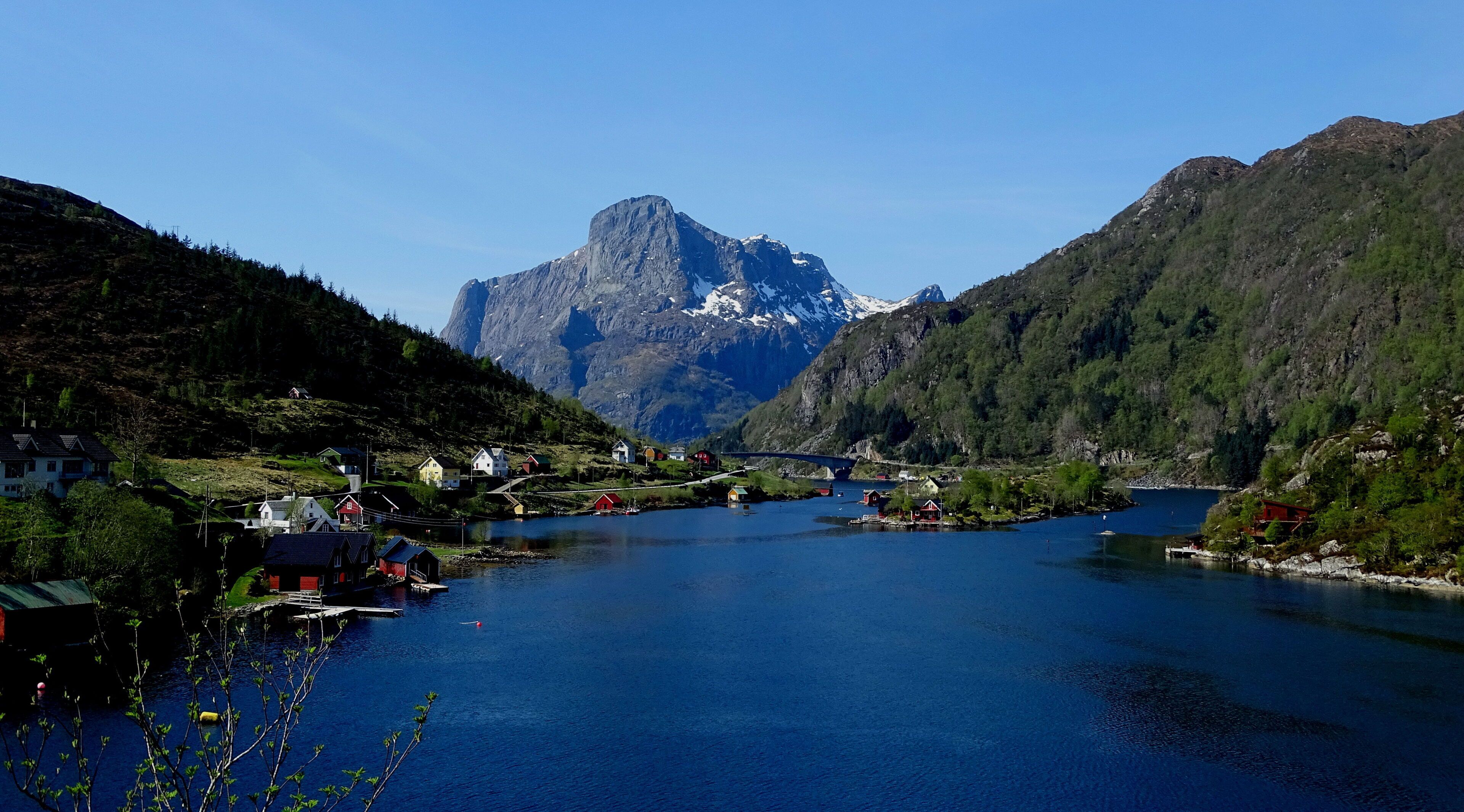 A serene lakeside village with houses along the water, a bridge connecting the areas, and mountains in the background.
