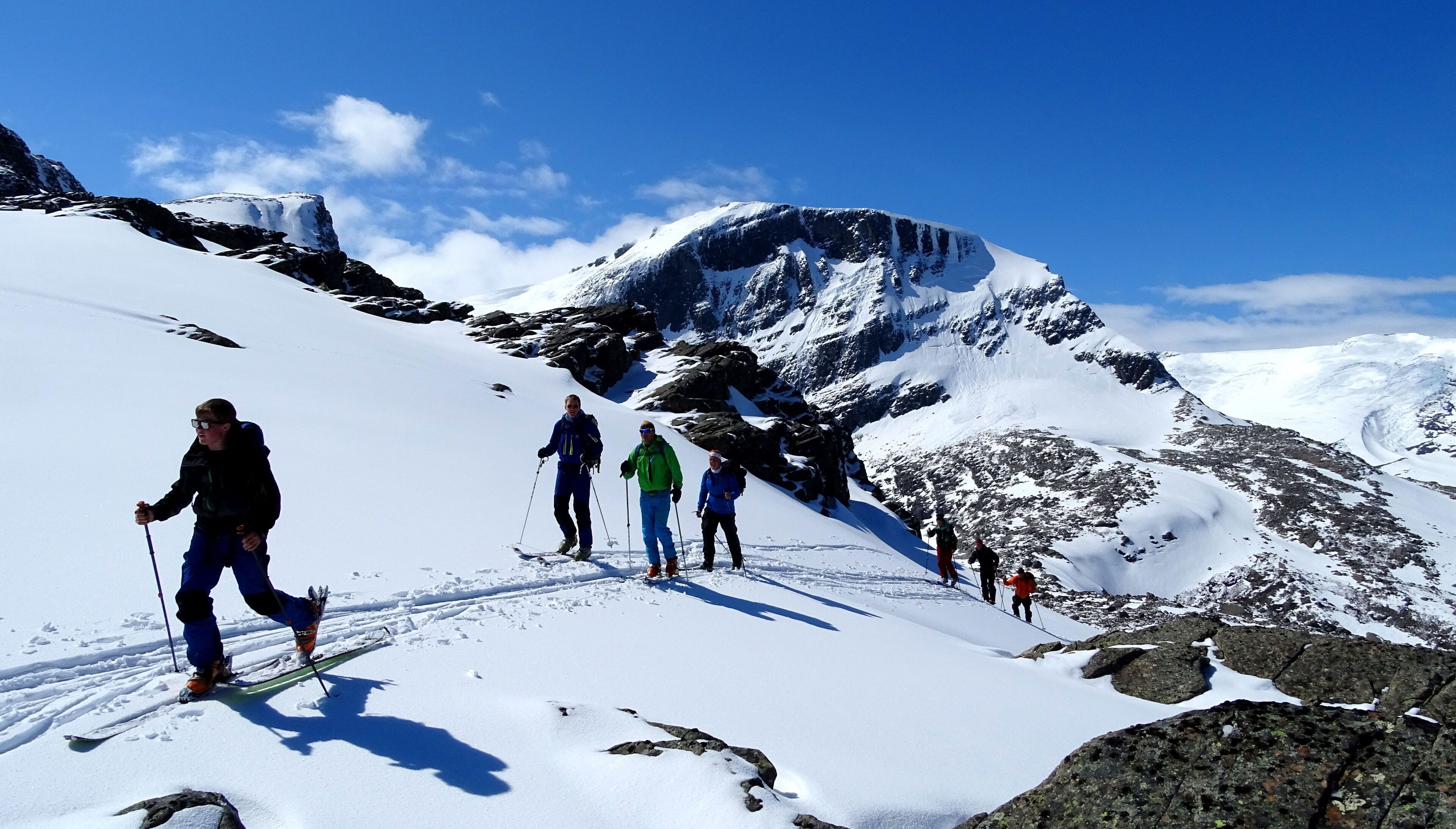 Un groupe de personnes fait de la randonnée sur un sentier de montagne enneigé, parsemé de rochers, sous un ciel dégagé.