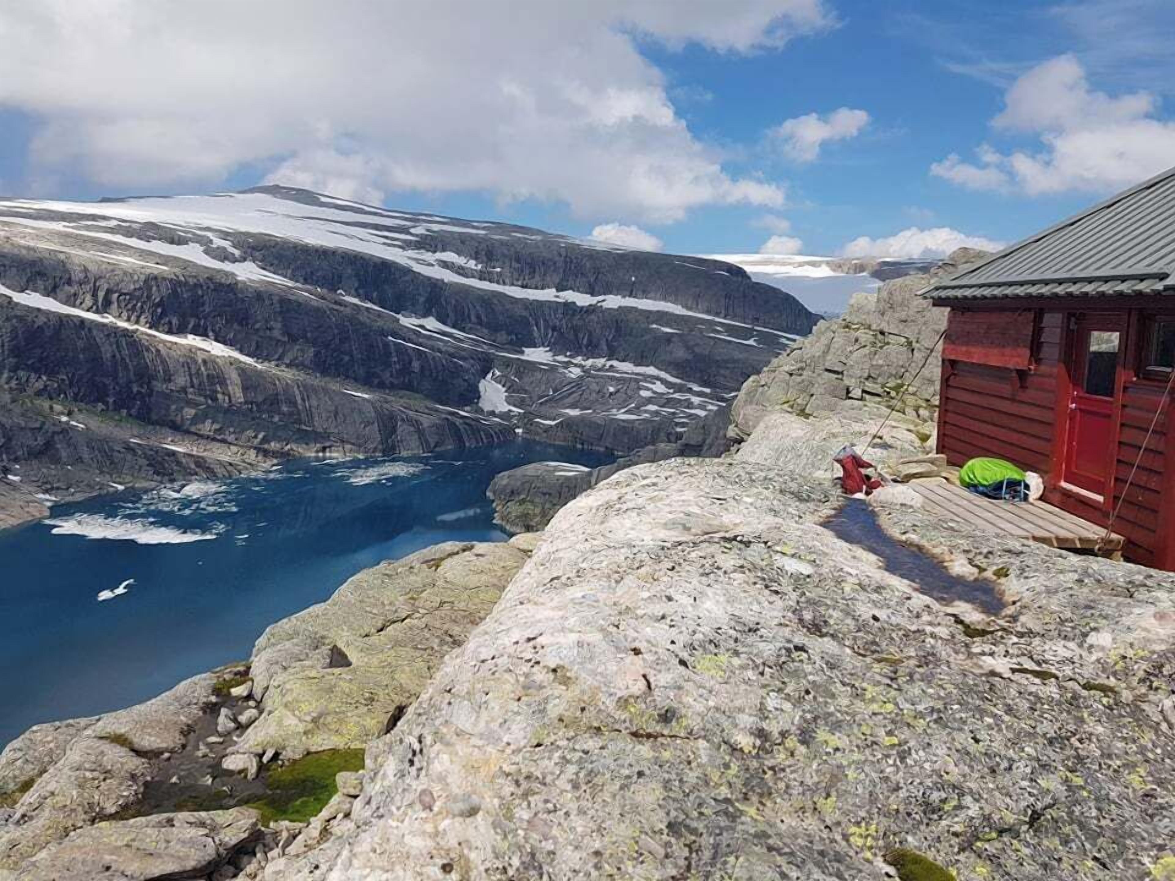 Ett kargt landskap med en liten stuga av trä som ligger på en klippavsats med utsikt över en sjö som får sitt vatten från en glaciär.