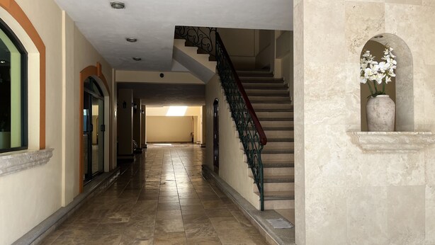 Un pasillo amplio con piso de mármol, una escalera con barandales ornamentales y un jarrón con flores blancas a la derecha.