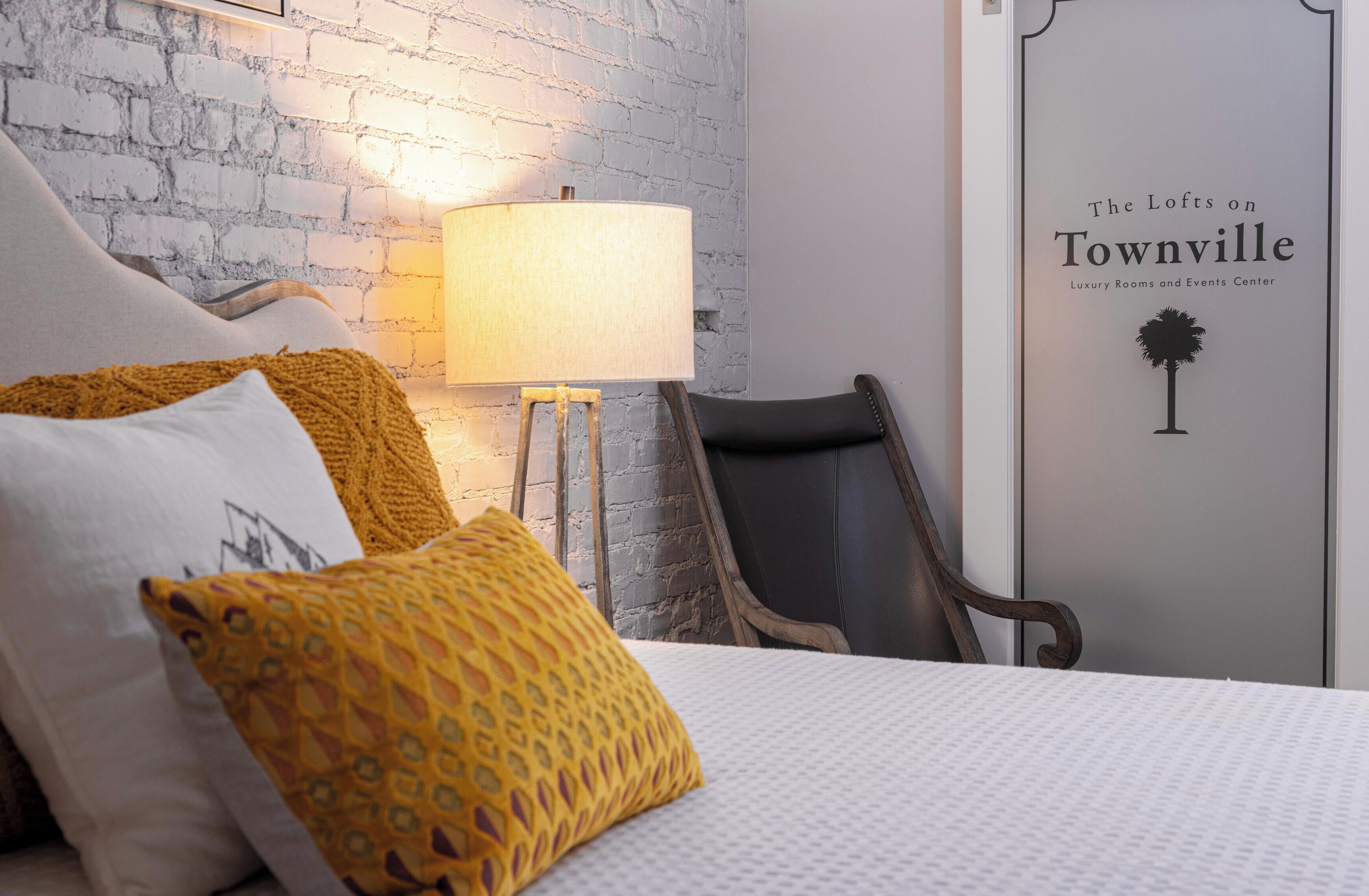 Una habitación de hotel con pared de ladrillo, una cama con almohadas estampadas, una lámpara de pie y una silla. La puerta tiene un letrero que dice 'The Lofts on Townville'.