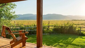 A wooden deck with a single lounge chair and a table, overlooking a green field with a fence and mountains in the distance. - Basecamp Stays: Upscale Retreat, Hot Tub, Mtn View (Victor)