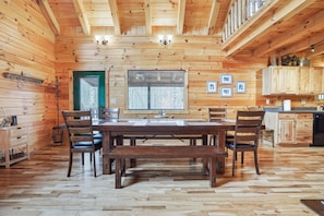 A wooden cabin interior with a dining table, chairs, a kitchen area, and a large window. - Applewood Lodge | Hocking Hills, Hot Tub, Fire Pit (Logan)