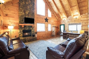 A log cabin interior with a stone fireplace, leather sofas, a dining area, and a TV mounted on the wall.