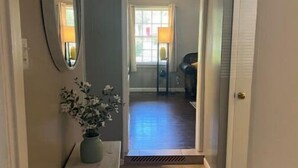 A hallway with a white console table, a potted plant, and a mirror on the left. The hallway leads to a room with a window, a lamp, and a chair. - Spacious 3BR House • Sleeps 8 • Near SGMC, VSU & Moody AFB • Parking (Valdosta)