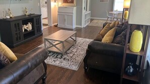 A living room with a dark leather sofa, a coffee table, and a TV mounted on the wall. There is a fireplace with a mantel and a painting above it. - Spacious 3BR House • Sleeps 8 • Near SGMC, VSU & Moody AFB • Parking (Valdosta)
