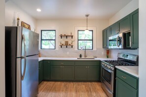A kitchen with green cabinets, a stainless steel refrigerator, a microwave, and a stove. There are two windows with plants on the windowsill.