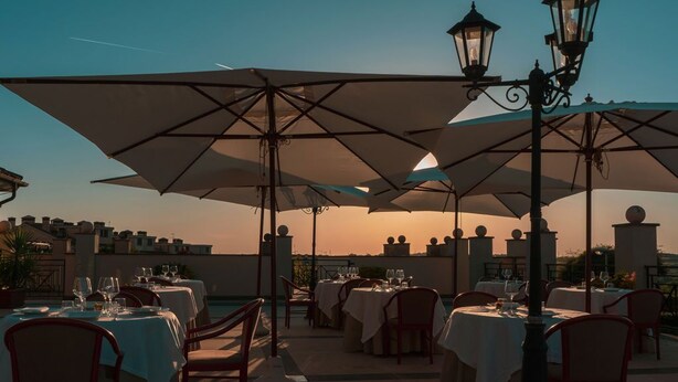 Un área de comedor al aire libre con mesas y sillas, sombrillas y farolas, con el atardecer de fondo.