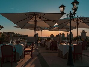 Un área de comedor al aire libre con mesas y sillas, sombrillas y farolas, con el atardecer de fondo.