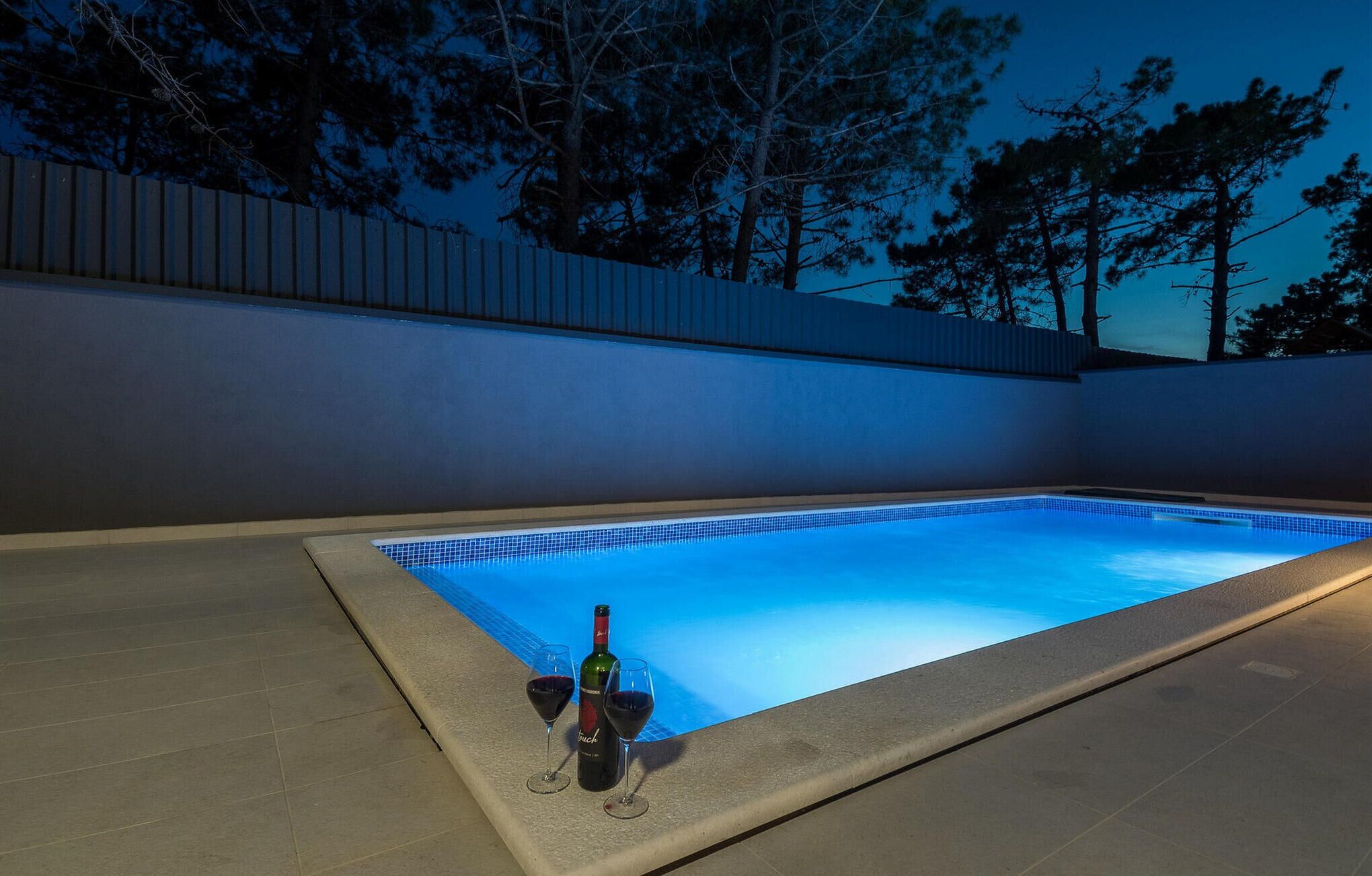 Une piscine dont le bord est recouvert de carrelage, une bouteille de vin et un verre posés sur le bord de la piscine.