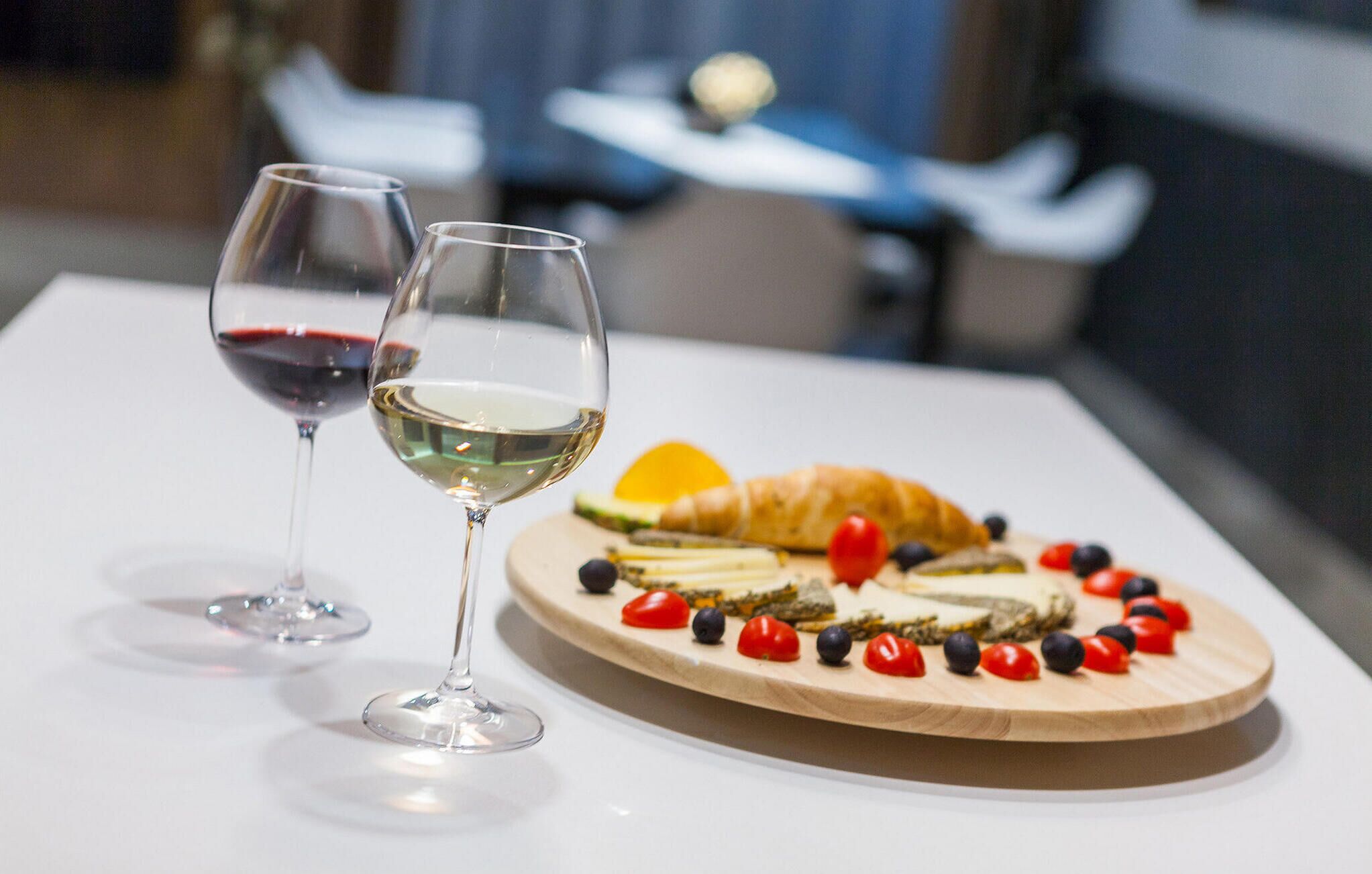 Dos copas de vino sobre una mesa con una tabla de madera que contiene un aperitivo de quesos y un croissant.