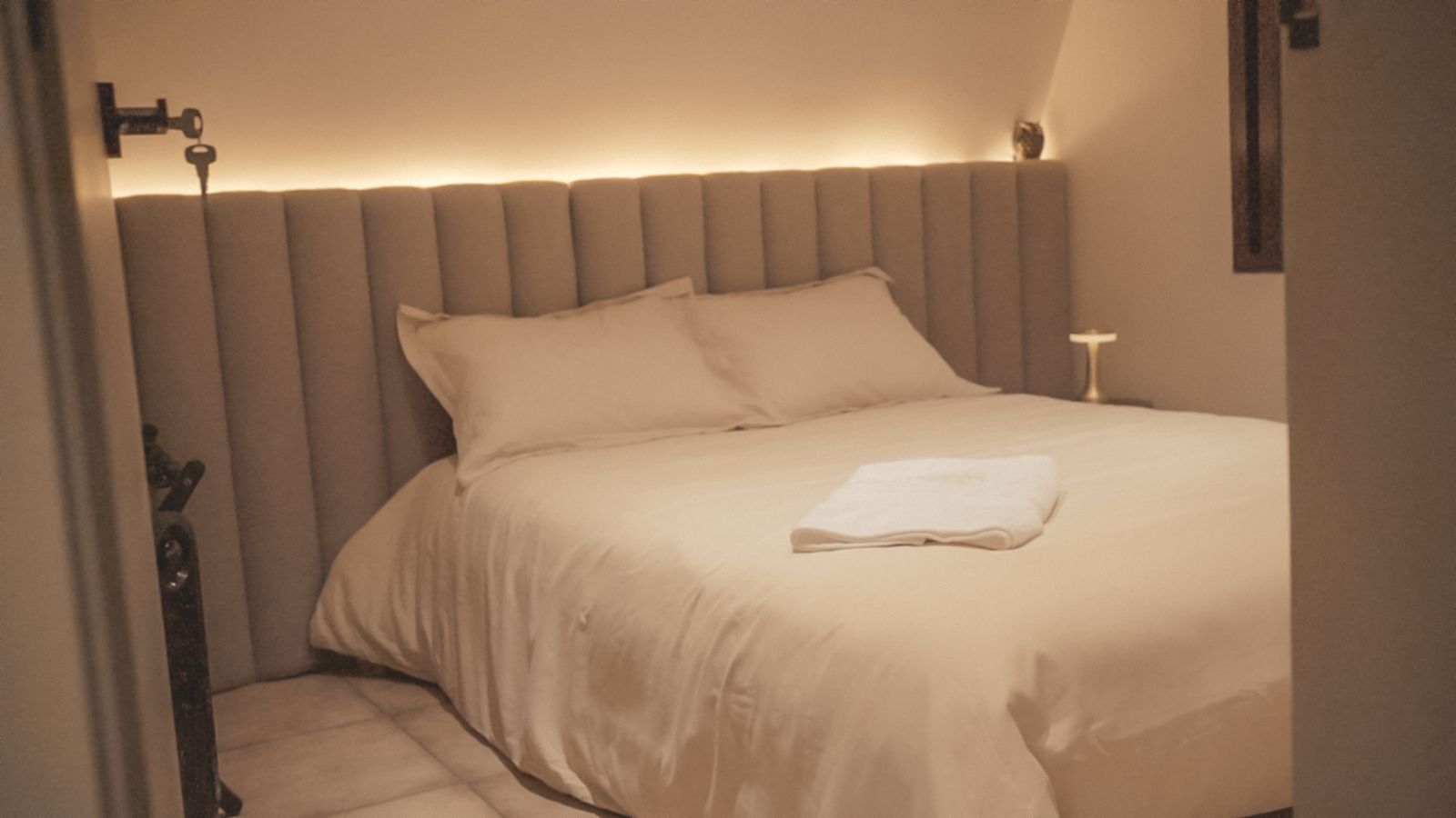 A neatly made bed with a folded white towel, a headboard, and a bedside table with a lamp.