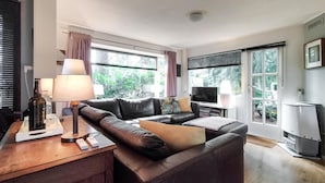 Un salon moderne avec un canapé en cuir, un téléviseur et une lampe. On y trouve des fenêtres avec des rideaux, une petite table avec une bouteille et une télécommande.