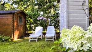 Un abri en bois, deux fauteuils de jardin blancs et un bouquet d’hortensias blancs dans un jardin, avec une maison en arrière-plan.