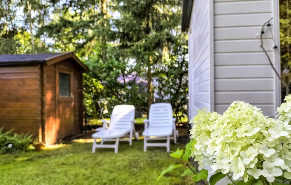 Un cobertizo de madera, dos sillones blancos y un grupo de flores de hortensia blancas en un patio trasero con una casa al fondo.