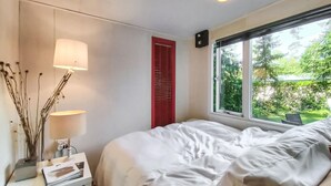 Une chambre à coucher avec un lit, des lampes de chevet, une petite table avec une lampe, une porte rouge et une fenêtre donnant sur un paysage verdoyant.