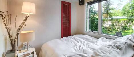 A bedroom with a bed, bedside lamps, a small table with a lamp, a red door, and a window with a view of greenery.