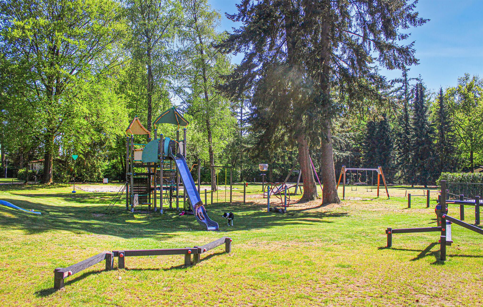 Un parc infantil amb tobogans i elements per escalar, envoltat d'arbres i zones enjardinades.