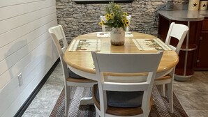 Een eetgedeelte met een ronde tafel, witte stoelen, een vaas met bloemen, een stenen muur, een raam, en een keuken met houten kasten en een aanrecht.