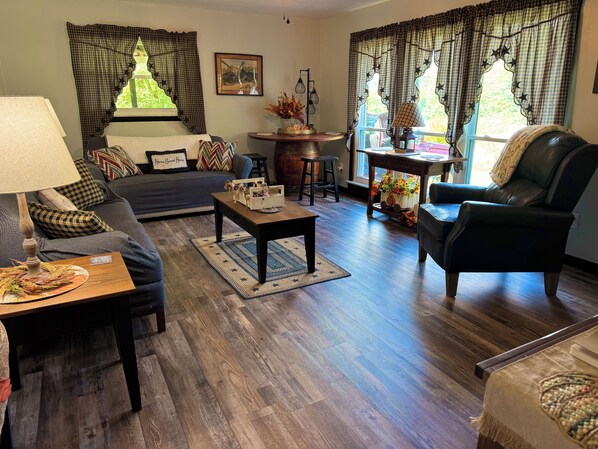A living room with a sofa, coffee table, and a chair. There is a window with curtains and a picture on the wall.