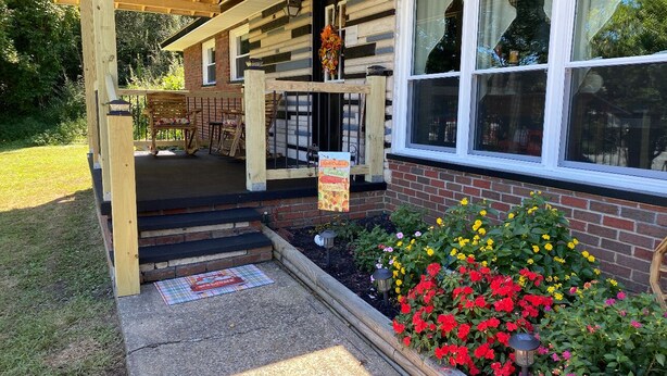A covered outdoor seating area with a wooden bench and a table, a brick building with large windows, and a flower bed with red and yellow flowers. - Beautiful Mountain View’s and Located on several acres for privacy (Chapmanville)