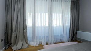 Un dormitorio con una cama, una ventana con cortinas blancas translúcidas y cortinas grises a los lados.