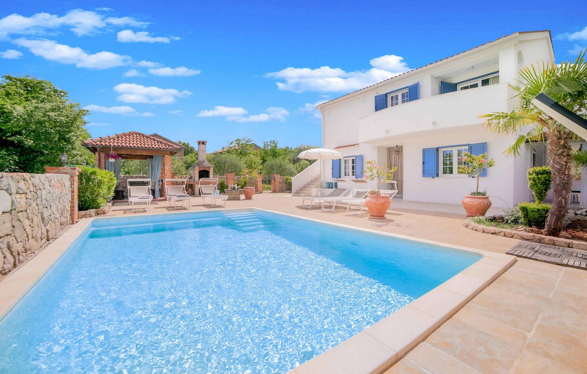 Ein Swimmingpool mit Liegestühlen und Grillplatz, umgeben von einer Steinmauer und Topfpflanzen.