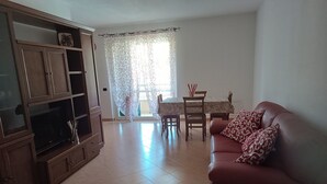 A living room with a brown leather sofa, a TV on a wooden cabinet, a dining table with chairs, and a window with patterned curtains. - Accogliente Appartamento a 3 km da Sestri Levante con Parcheggio Privato (Casarza Ligure)