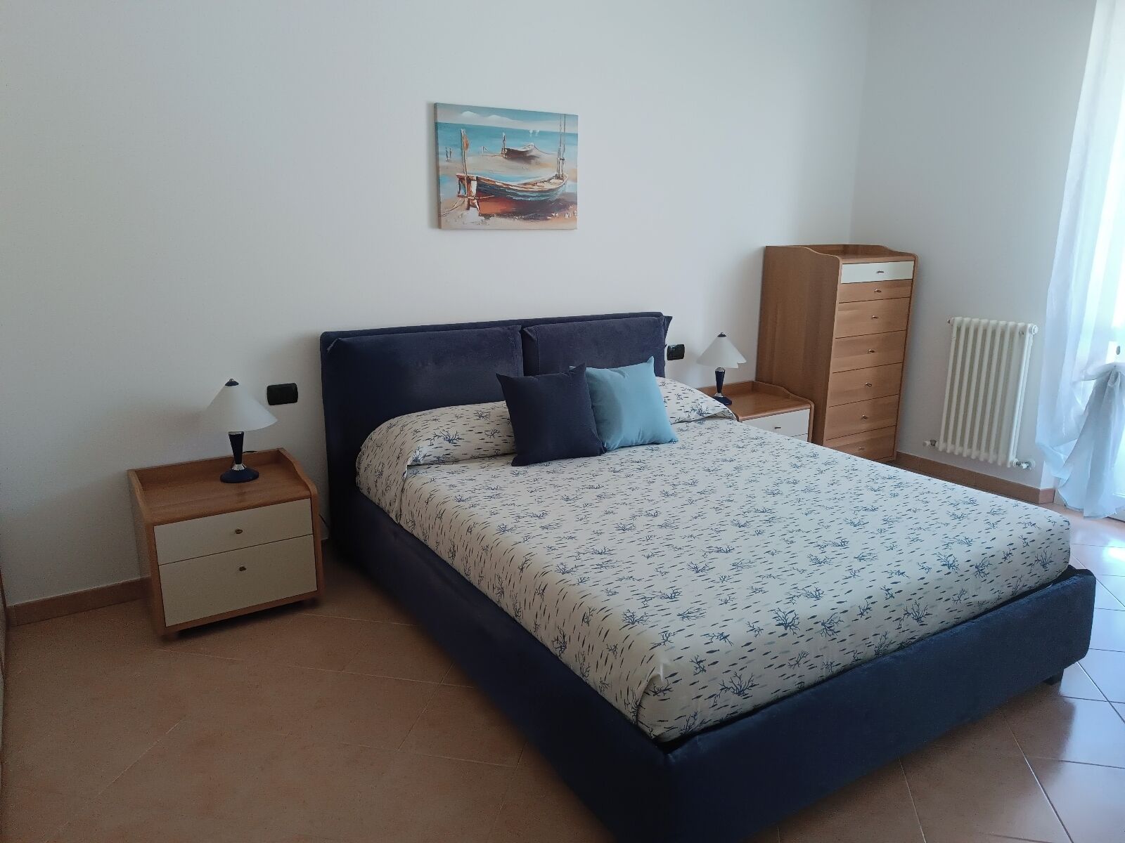 A bedroom with a bed, nightstands, a dresser, a painting, and a radiator.