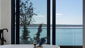 A bathroom with a bathtub, a glass of wine, and a bottle of sunscreen by the window overlooking a body of water. - Villa Beatrice (Soho) (Starigrad)