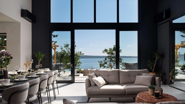 A modern living room with a large window overlooking a body of water, a sofa, and a dining area with chairs and a table set for a meal. - Villa Beatrice (Soho) (Starigrad)