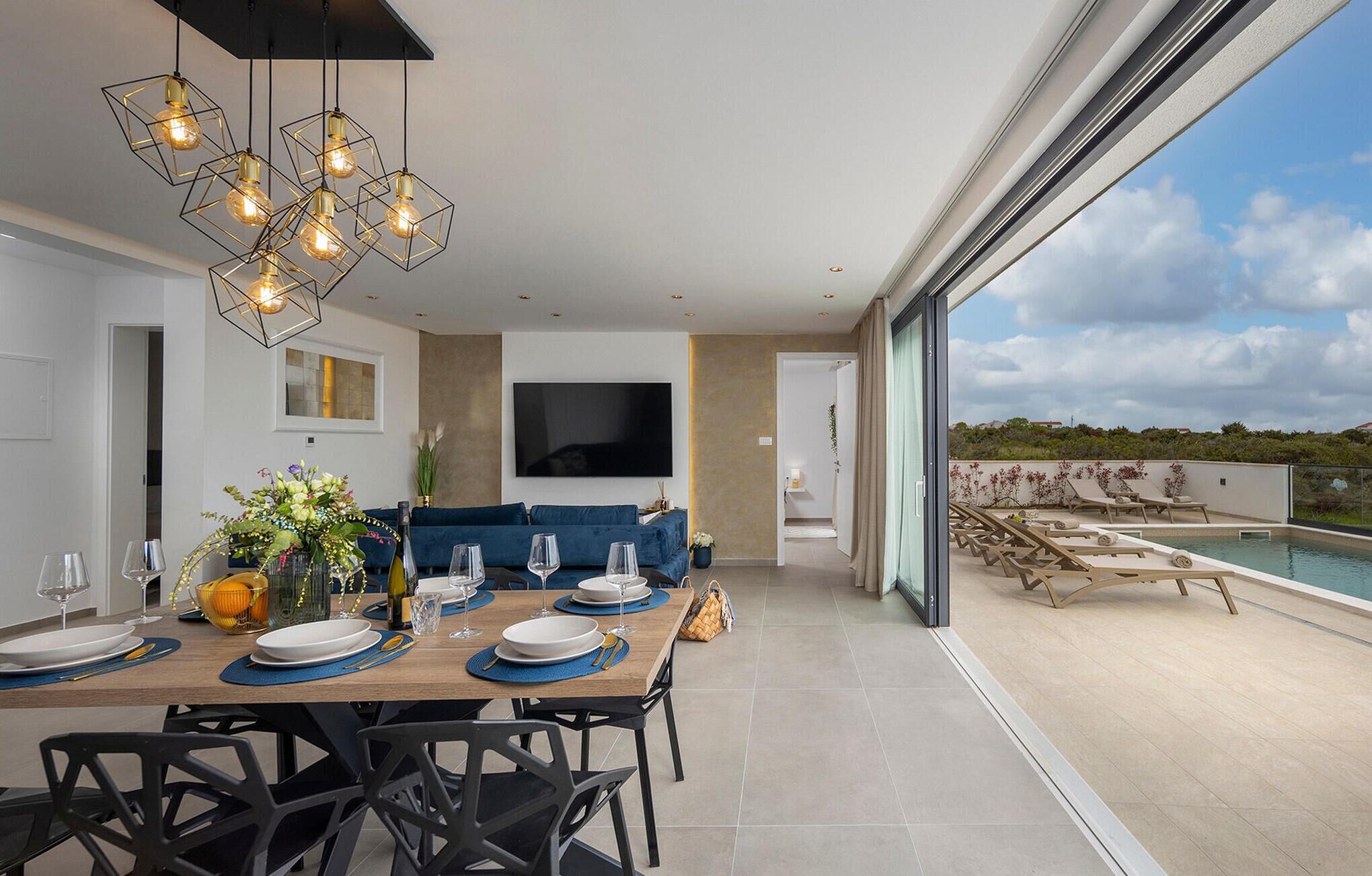 A modern dining area with a table set for four, a TV mounted on the wall, and a view of a pool and landscape.