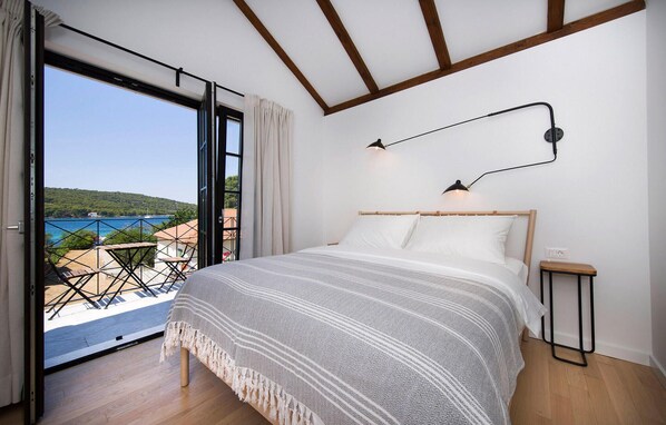 A bedroom with a large bed, a nightstand, a wall-mounted lamp, and a view of a balcony with a table and chairs.