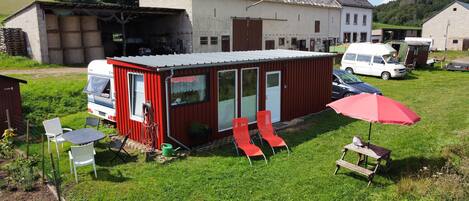 Una pequeña casa rodante roja con garaje, una furgoneta blanca y una mesa de picnic con sombrilla roja en un césped.