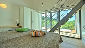 Une chambre moderne avec un grand lit, un oreiller vert et un oreiller à rayures. Un escalier mène à un balcon d’où l’on peut admirer la verdure.