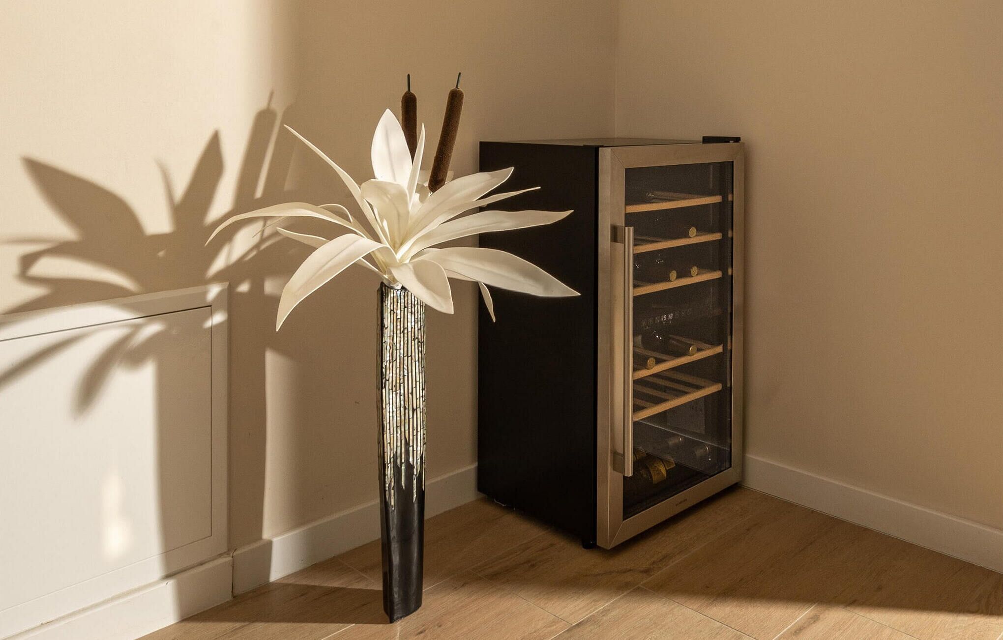 Une pièce d’angle avec un grand vase rempli de fleurs blanches, un meuble noir muni d’un porte-bouteilles et un plancher en bois.