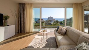 Ein modernes Wohnzimmer mit einer Couch, einem kleinen Tisch und einem Fernseher auf einem Schrank. Große Fenster bieten Blick auf das Meer und Gebäude.