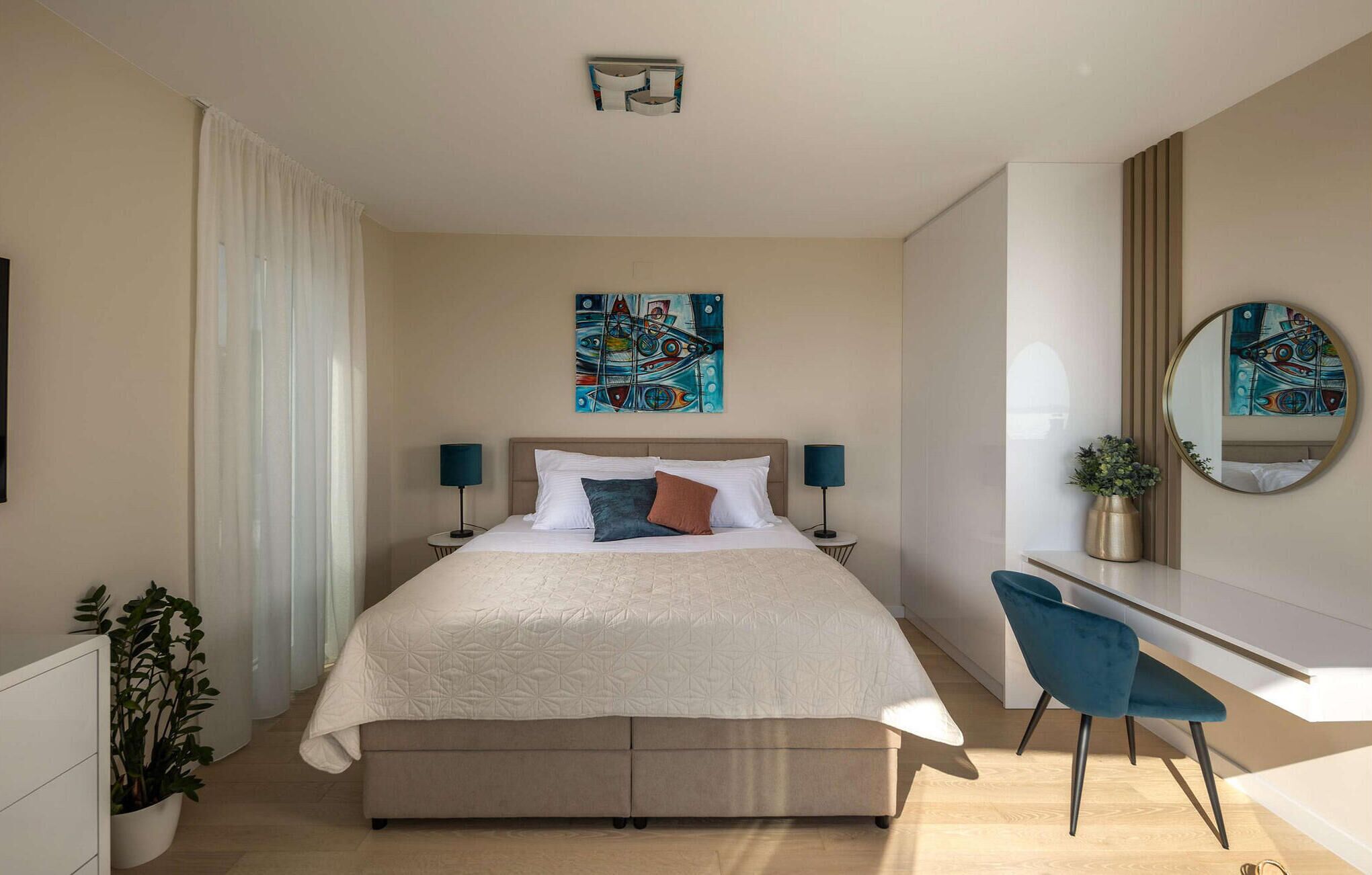 Quarto de hotel moderno com uma cama grande, abajures, uma mesa de trabalho com cadeira, uma planta em vaso e obras de arte nas paredes.