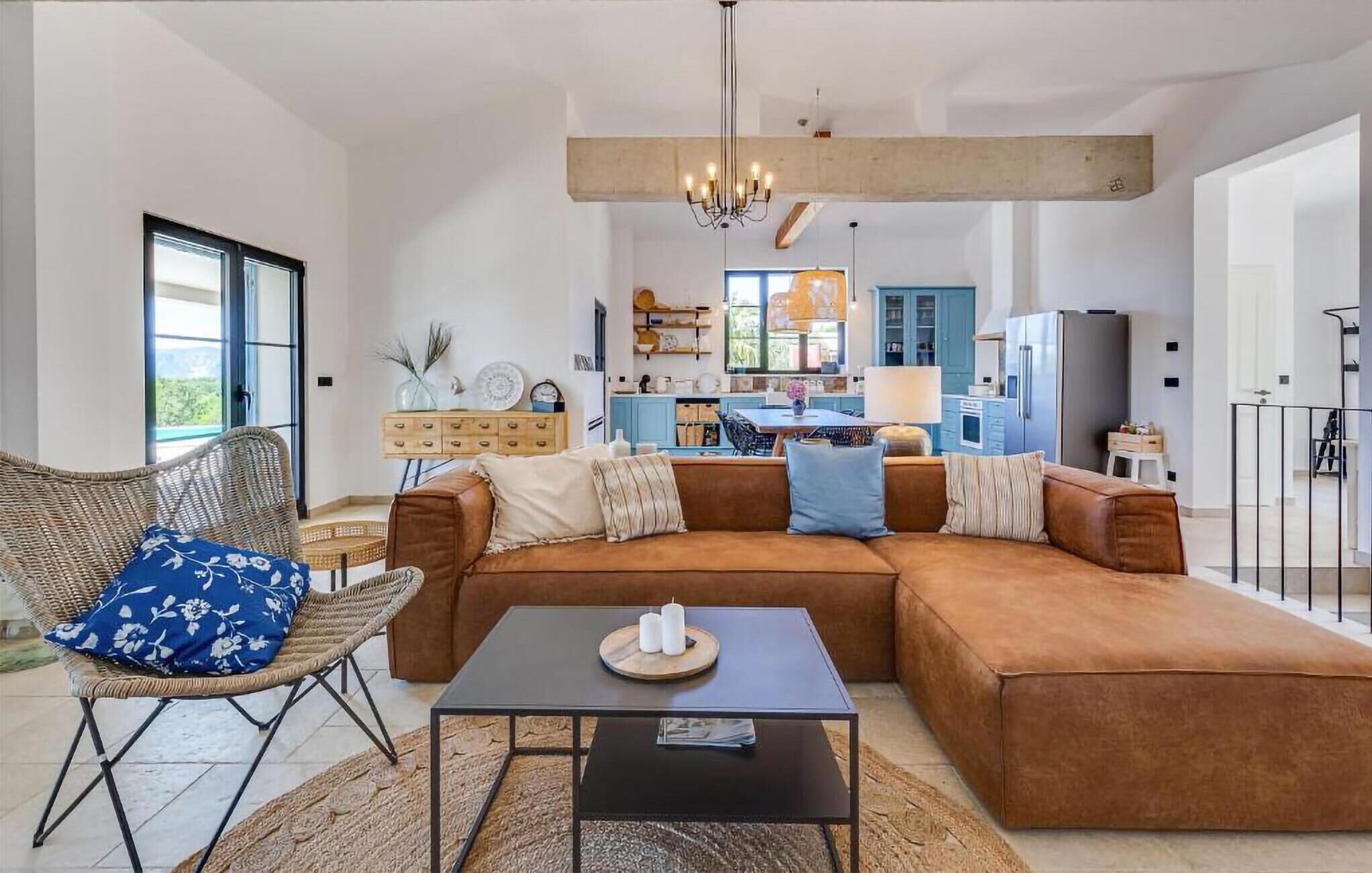 A spacious living room with a large brown sectional sofa, a wicker chair, and a black coffee table. A kitchen with blue cabinets and a hanging light fixture is visible in the background.