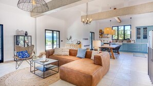 Amplia sala de estar con un sofá seccional marrón grande, un sillón de mimbre y una mesa de comedor con sillas. Hay una zona de cocina con armarios azules y una lámpara colgante.