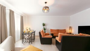 Un salon moderne comprenant un canapé, un fauteuil et un pouf, une table à manger avec des chaises et un téléviseur.