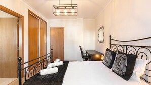 Un lit bien fait, recouvert d’une couverture sombre, une porte en bois, un bureau avec une chaise et un miroir accroché au mur.