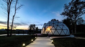 Una estructura de domo geodésico con un sendero pavimentado que conduce a él, rodeado de árboles y una masa de agua al fondo.
