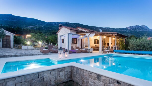 A poolside area with a stone wall, lounge chairs, and a small building with a tiled roof.