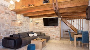 A cozy living room with a stone wall, a wooden ceiling, a black sofa, a wooden coffee table, and a blue ottoman.
