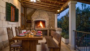 An outdoor patio with a stone fireplace, wooden furniture, and a view of trees.