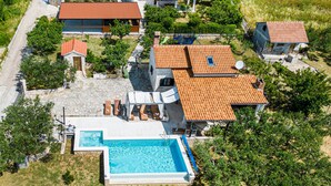 An aerial view of a residential area with a swimming pool, multiple houses with red-tiled roofs, and surrounding greenery.