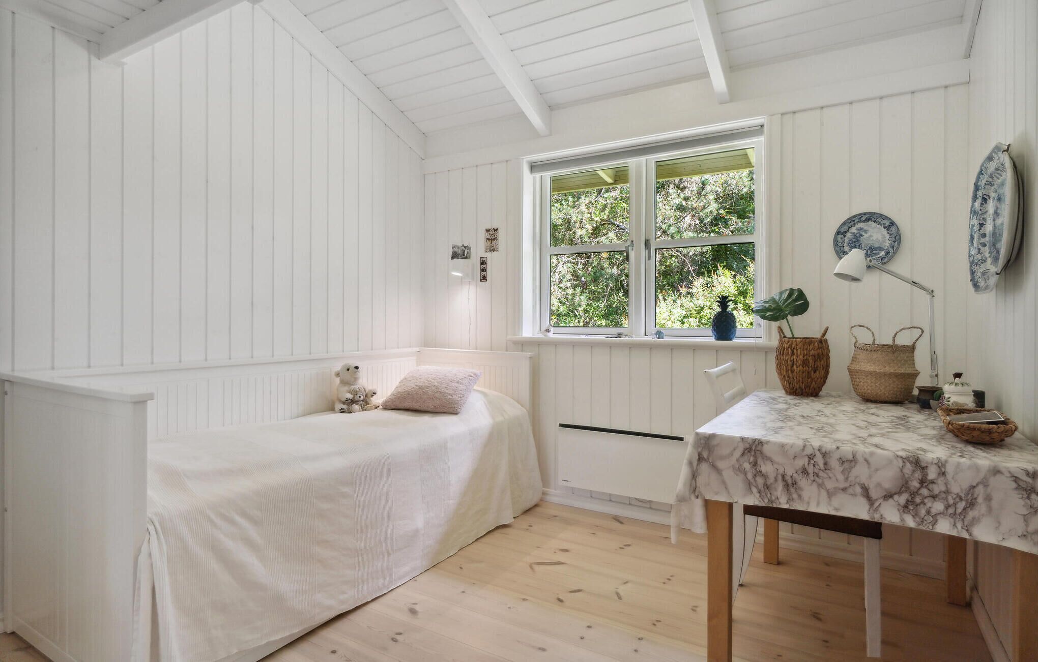 Een kleine kamer met witte wanden, een enkel bed, een raam met uitzicht op groen, een tafel met een marmeren blad en een lamp.