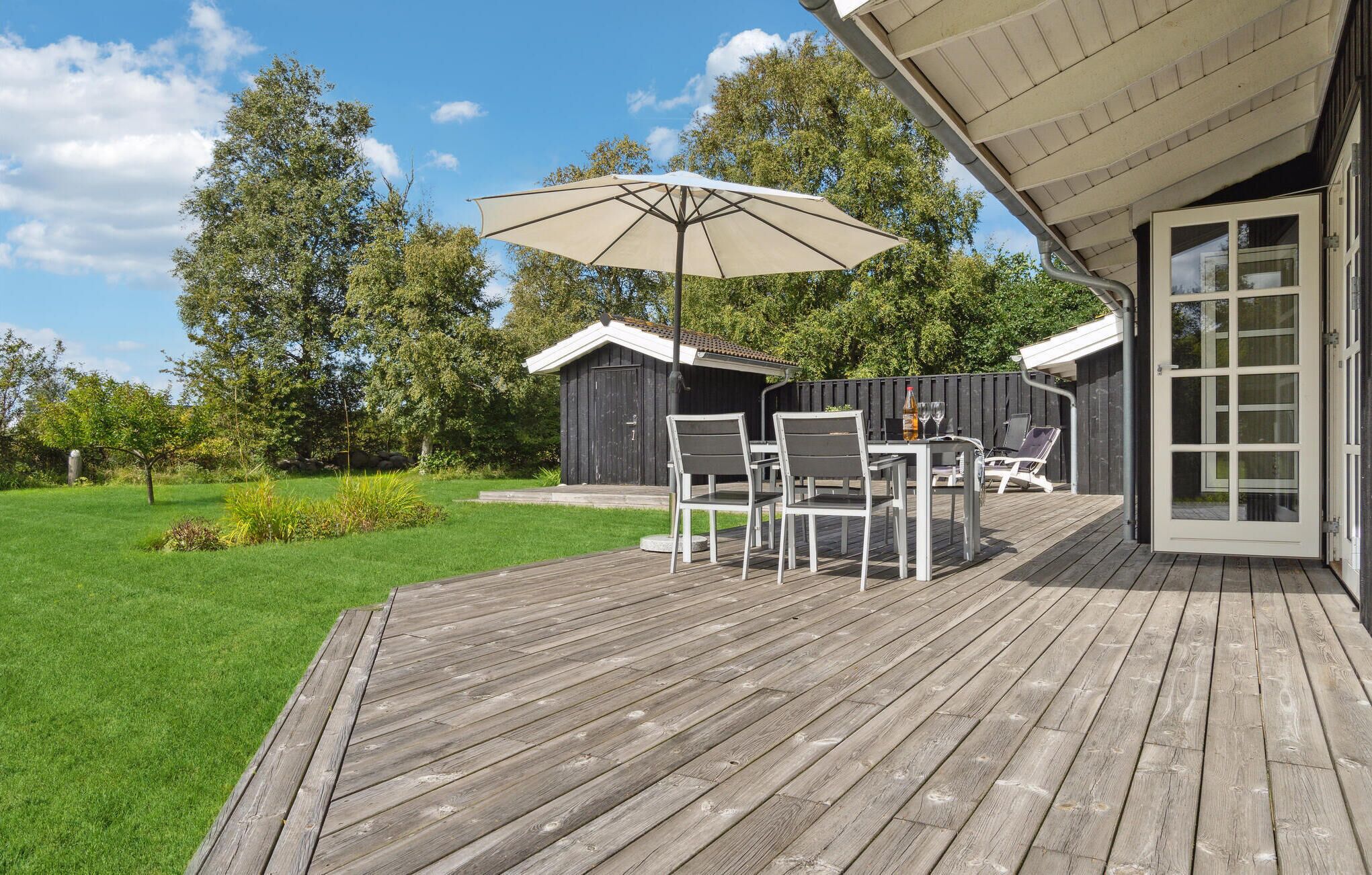 Eine Holzterrasse mit Gartenmöbeln, einem großen Sonnenschirm und einem Gebäude mit Glastür.