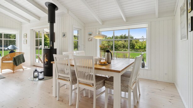 A cozy room with a wooden table, white chairs, a black stove, and large windows offering a view of greenery. - Gorgeous home in Jerup with sauna (Jerup)